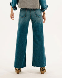 A person is shown from the back against a white background, wearing ASKK NY’s Mercer Jean in Gunnison—a high rise, wide-leg blue Japanese denim—paired with a fringed-edge gray top and leopard-print platform shoes, arms relaxed at their sides.