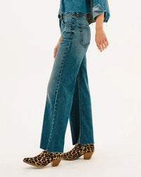 A person wearing ASKK NY’s Mercer Jean in Gunnison, a denim shirt, and leopard-print heeled ankle boots stands against a plain white background.