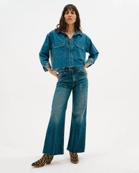 A woman with long brown hair and bangs poses with hands on hips in ASKK NY’s Mercer Jean in Gunnison, a high-rise wide-leg jean, styled with a blue Japanese denim button-up shirt and leopard print ankle boots against a white background.