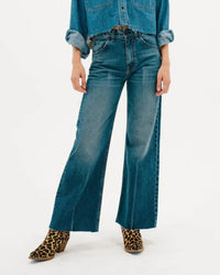 A person wears the Mercer Jean in Gunnison by ASKK NY—a high-rise, wide-leg style—paired with a blue denim shirt and leopard print ankle boots, standing against a plain white background.
