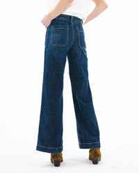 A person is shown from behind against a white background wearing ASKK NY’s Sailor in Dark Stone high-rise wide-leg Japanese denim jeans and brown heeled shoes.