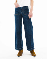 A person wearing a white shirt, ASKK NY Sailor in Dark Stone high rise wide leg jeans with front patch pockets, and brown suede ankle boots, stands against a plain white background.