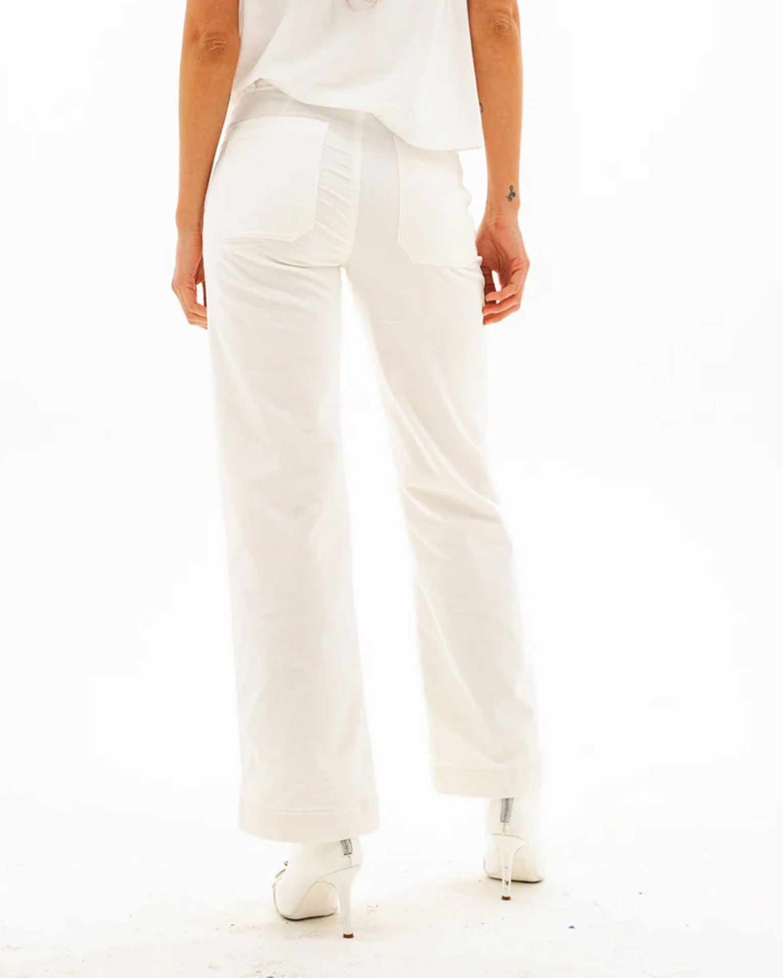 A person stands facing away, wearing ASKK NY Sailor Corduroy in Eggshell—white high-rise pants with two back pockets, one with ruching—paired with a white top and heels against a plain white background.