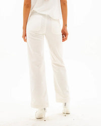 A person stands facing away, wearing ASKK NY Sailor Corduroy in Eggshell—white high-rise pants with two back pockets, one with ruching—paired with a white top and heels against a plain white background.