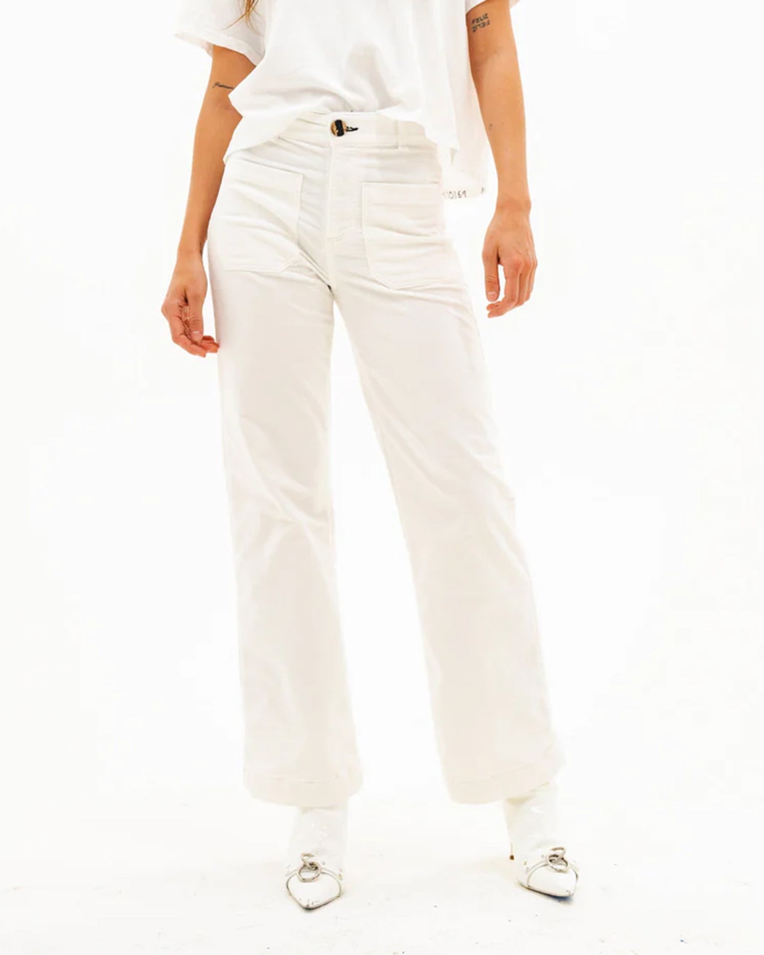 A person, visible from the waist down, stands against a plain white background wearing ASKK NY’s Sailor Corduroy in Eggshell and white shoes with silver buckles.