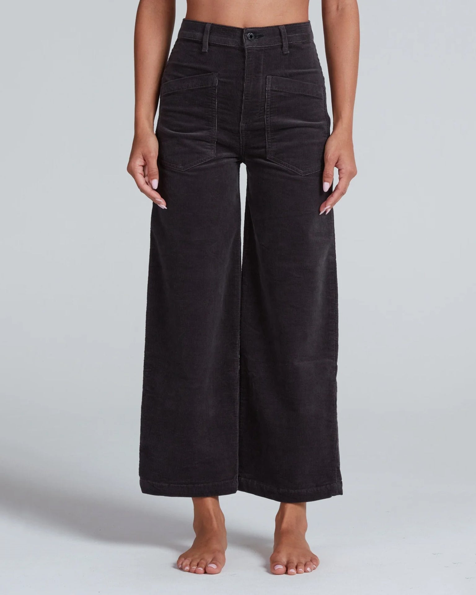 A person stands barefoot against a plain light gray background wearing ASKK NY’s Virginia Corduroy in Charcoal—high-rise, wide-leg pants featuring front patch pockets, visible stitching, and a button fly.