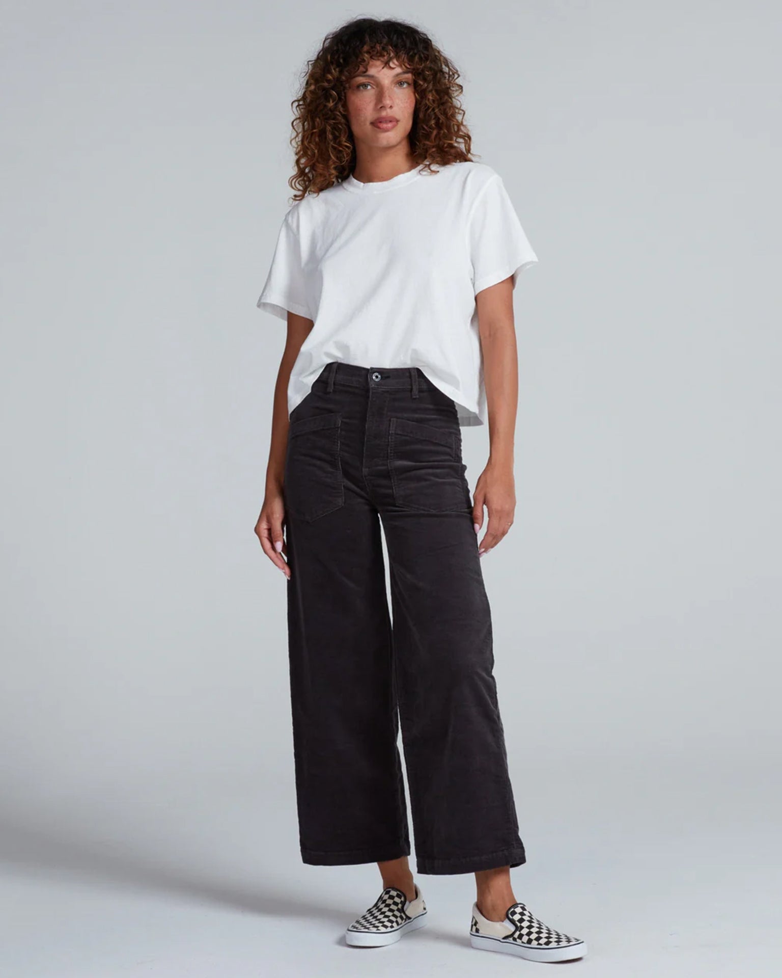 A person with curly hair wears a loose white T-shirt, ASKK NY’s Virginia Corduroy in Charcoal high-rise wide-leg pants, and checkered slip-on shoes while standing against a plain light gray background.