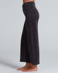 A person stands barefoot in profile against a plain light gray background, wearing ASKK NY’s Virginia Corduroy in Charcoal—high-rise, wide-leg pants with front patch pockets and a cropped length.
