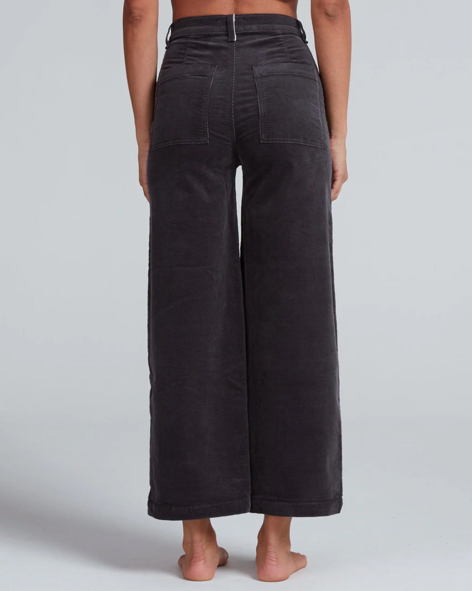 A person stands barefoot, shown from the back, wearing ASKK NY Virginia Corduroy wide-leg, high-rise pants in Charcoal with visible back pockets and seam details. The cropped pants contrast against a plain light gray background.
