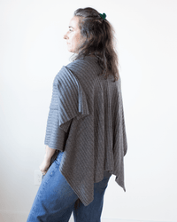 A person with wavy brown hair wears the Autumn Cashmere Rib Drape Poncho in Flannel and blue jeans, standing turned slightly away against a plain white background.