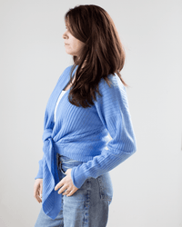 A woman with long brown hair stands in profile, wearing the Autumn Cashmere Tie Front Rib Cardigan in Cielo over a white top and blue jeans, set against a plain white background.