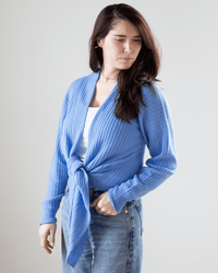 A woman with long brown hair wears the Tie Front Rib Cardigan in Cielo by Autumn Cashmere over a white top and blue jeans, standing against a plain light background and gazing slightly to the side.