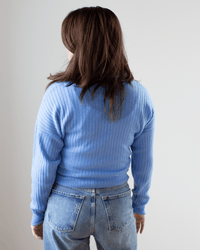 A person with long brown hair stands facing away from the camera against a plain light background, wearing Autumn Cashmere’s Tie Front Rib Cardigan in Cielo and blue jeans.