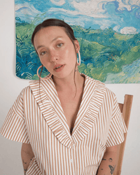 A light-skinned woman in a striped, ruffled-collar shirt sits on a wooden chair, wearing B&L's C Hoop in Large in Runaway Bunny—mango wood earrings with 14k gold filled posts—glistening as she poses before green and blue fields.
