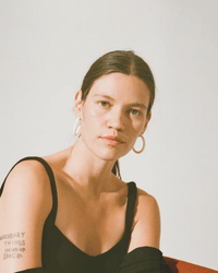 A woman with straight brown hair, wearing B&L's C Hoop Small earrings in Chanel Ivory and a black tank top, sits against a plain light background. She has an arm tattoo that reads “NECESSARY THINGS TAKING UP SPACE.”.