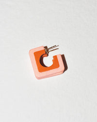 The B&L Square Hoop in Helen earring features a pink square C design with an orange inner border, displayed on a white textured surface—a stylish piece of wearable art jewelry.
