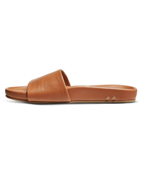 The Gallito in Tan by beek. is a brown leather slide sandal with a wide single strap, flat sole, and molded footbed, shown from the side on a white background. Two small metallic rivets are visible near the heel.