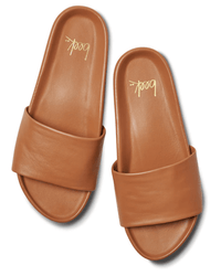 A pair of Gallito in Tan leather slides by beek., with wide straps, flat soles, and a molded footbed, displayed side by side on a white background. The brand name “beek..” is printed in gold on the insoles.