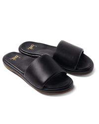 The Puffbird in Black by beek.. are black slide sandals with wide straps and flat soles, displayed at an angle on a white background. Their insoles show the gold beek.. logo and provide molded arch support for comfort.