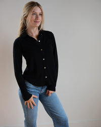 A woman with long blonde hair wears the Bella Dahl Button Up Knit Top in Black and light blue jeans, standing against a plain light-colored wall, smiling slightly and looking off to the side.