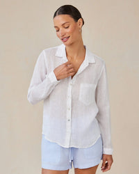 A woman wears the Bella Dahl Pocket Button Down in White and light blue shorts, standing against a plain beige background, smiling and looking down with one hand touching her collar.