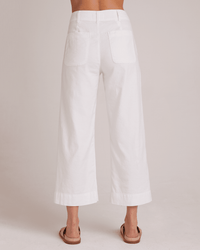 A person stands barefoot, seen from behind, wearing Bella Dahl's Saige Wide Leg Crop in White—high-rise linen-cotton blend trousers with large square back pockets and a cuffed hem—against a plain light background.