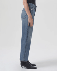 A person stands in profile wearing AGOLDE’s 90s Crop Mid Rise Loose Straight in Hooked, featuring straight-leg jeans with raw hems and black pointed-toe ankle boots, against a plain light background.