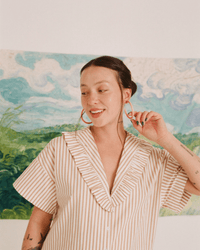 A smiling woman with dark hair in a bun wears B&L's C Hoop in Large from Charlotte's Hoop, along with a striped blouse. She stands before a vibrant landscape backdrop featuring green fields and a blue sky.