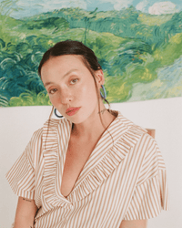 A woman with light skin and brown hair wears a striped, ruffled top and B&L's C Hoop in Small in Blueberries for Sal earrings, sitting before a vibrant green abstract landscape painting. She gazes calmly at the camera.