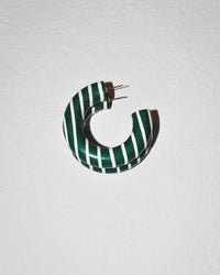 The Small Chunk Hoop in Elaine by B&L is a chunky, circular mango wood earring with white and dark green diagonal stripes, a small gap, and two metal prongs for fastening, shown on a textured white surface.