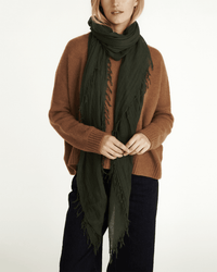A person wears a brown sweater, dark wide-leg pants, and a Chan Luu Cashmere & Silk Scarf in Kombu Green wrapped around their neck, standing against a plain light background.
