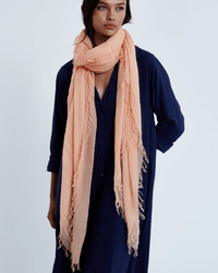 A woman wears a loose, long-sleeved navy dress and a Chan Luu Cashmere & Silk Scarf in Peach wrapped around her neck, standing against a plain light background.