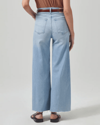 Person wearing the Lyra Crop Wide Leg in Marquee jeans by Citizens of Humanity with frayed hems, a brown belt, a brown top, and black sandals, standing with their back to the camera against a plain light background.