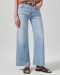 A person wearing Citizens of Humanity Lyra Crop Wide Leg in Marquee jeans, a brown belt, brown top, and brown sandals stands against a plain light background.