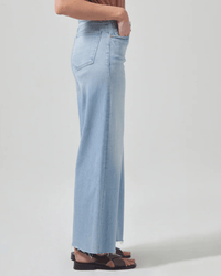 A person stands in profile wearing Citizens of Humanity Lyra Crop Wide Leg in Marquee, light blue jeans with raw hems and black sandals, set against a plain light background.
