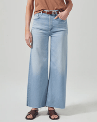 A person wears Citizens of Humanity Lyra Crop Wide Leg in Marquee jeans with a brown belt, brown top, and dark brown sandals. They stand against a plain light background with one hand in their pocket.