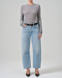 A person wears a fitted gray sweater, Citizens of Humanity Miro Relaxed Jean in Gemini—a light blue, barrel-leg style—and black pointed-toe heels, standing against a plain light background with hands in the jean’s front pockets.