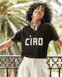 A curly-haired woman smiles with her eyes closed, wearing the Clare V. Block Ciao Original Tee in Black w/ Cream, cream trousers, hoop earrings, and a patterned neck scarf, standing outdoors by a railing with palm trees behind her.