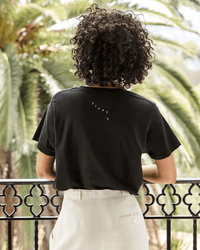 Seen from behind on a balcony with palm trees, a person with curly hair wears Clare V.'s Block Ciao Original Tee in Black w/ Cream and light high-waisted pants for an effortlessly cool look.