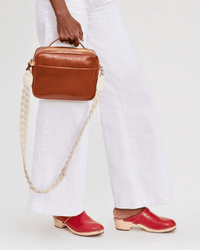 A person holds a Clare V. Braided Rope Crossbody Strap in Cream with Leather Tabs, paired with white wide-leg pants and red clogs against a neutral background.