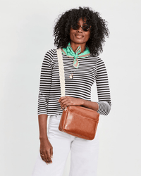 A person smiles against a plain light background, wearing sunglasses, a striped shirt, white pants, a green scarf, and carrying a Clare V. Braided Rope Crossbody Strap in Cream with leather tabs on their brown leather crossbody bag.