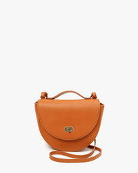 The Clare V. Elodie in Cuoio is a small, round bag made from Italian vachetta leather with a flap closure, top handle, and long adjustable strap, shown against a plain white background.