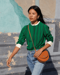 A woman with long dark hair walks outdoors against a painted wall, wearing a green and black striped sweater, light blue jeans, and carrying the Clare V. Elodie in Cuoio Italian vachetta leather crossbody handbag.