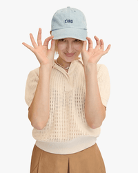 A person smiles playfully while holding the brim of the Clare V. Emb Petit Block Ciao Baseball Hat in Light Denim with Navy, styled with a short-sleeved cream knit top and tan skirt against a plain white background.