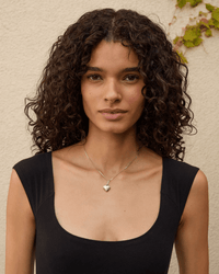 A woman with curly dark hair wears a black sleeveless top and the Clare V. Fine Box Chain Necklace 16in in Sterling Silver, standing before a beige wall with a small vine in the top right corner.