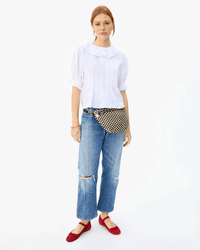 A woman stands facing forward against a plain white background, wearing a white ruffled blouse, blue ripped jeans, red shoes, and the Clare V. Grande Fanny in Black & Cream Woven Checker.