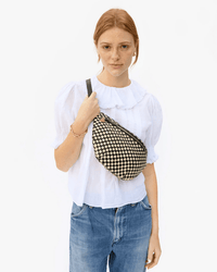 A woman with red hair, in a white blouse and blue jeans, stands against a plain background. She carries the Clare V. Grande Fanny in Black & Cream Woven Checker over her shoulder and looks directly at the camera.