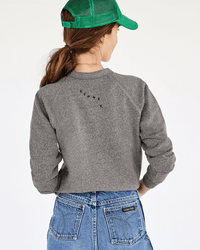 A woman with long brown hair, wearing a green cap, blue jeans, and the Clare V. Masculin Feminin Sweatshirt in Grey w/ Black, stands with her back to the camera against a plain, light background.
