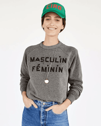 A person smiles while wearing a Clare V. Masculin Feminin Sweatshirt in Grey w/ Black, blue jeans, a green cap with red lettering, and a necklace, standing against a plain white background.