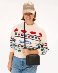 A smiling person in a beige baseball cap, colorful heart and fish sweater, blue jeans, and the Clare V. Midi Sac in Black Napetto stands against a plain background.